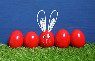 One egg with drawn face and ears as Easter bunny among others on green grass against blue wooden background