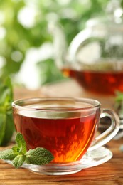 Photo of Glass cup of aromatic black tea with fresh mint on wooden table against blurred background. Space for text