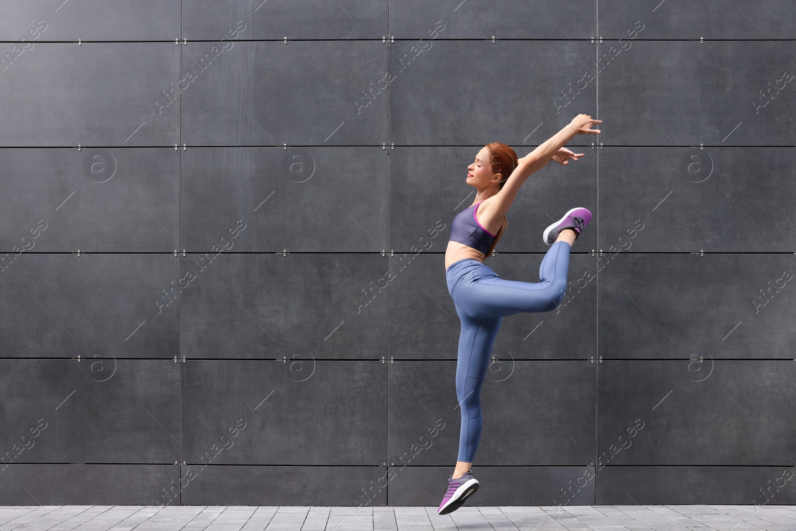 Photo of Beautiful woman in gym clothes jumping near dark grey wall on street, space for text