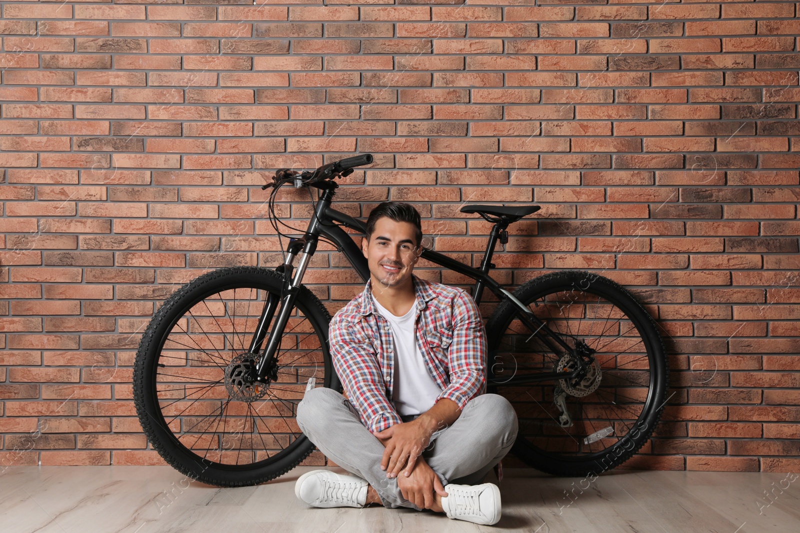 Photo of Handsome young man with modern bicycle near brick wall indoors