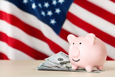 Photo of Piggy bank and dollar banknotes on white wooden table against American flag, space for text