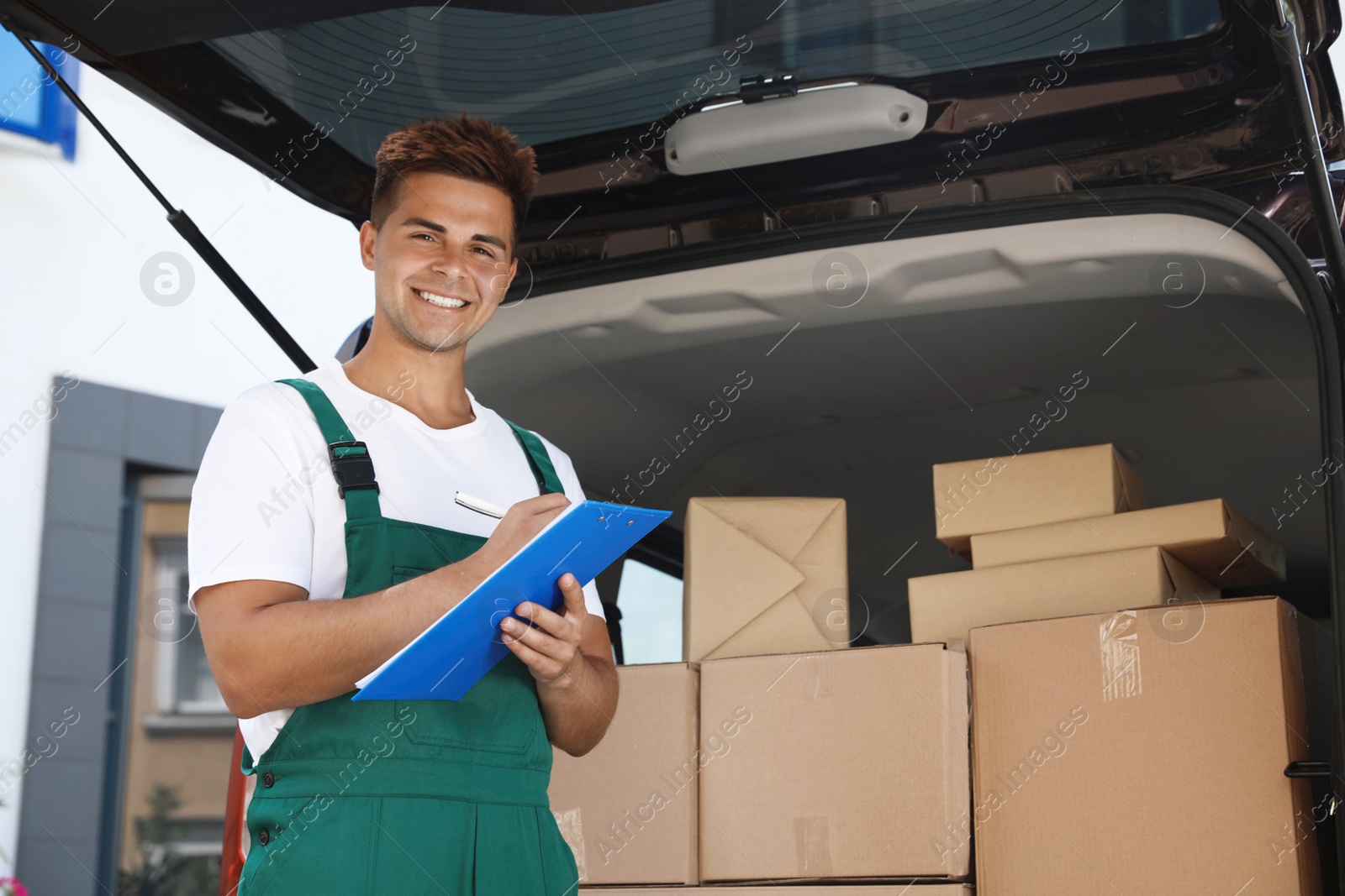 Photo of Young courier holding clipboard near delivery van with parcels outdoors