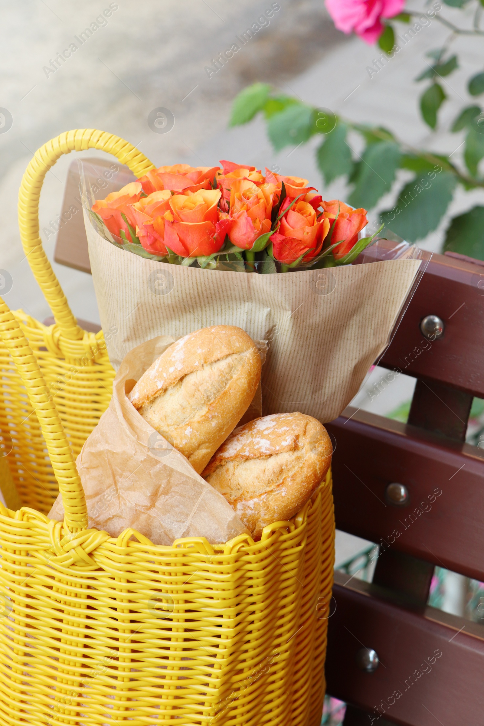 Photo of Beautiful bouquet of roses and baguettes in yellow wicker bag on bench outdoors