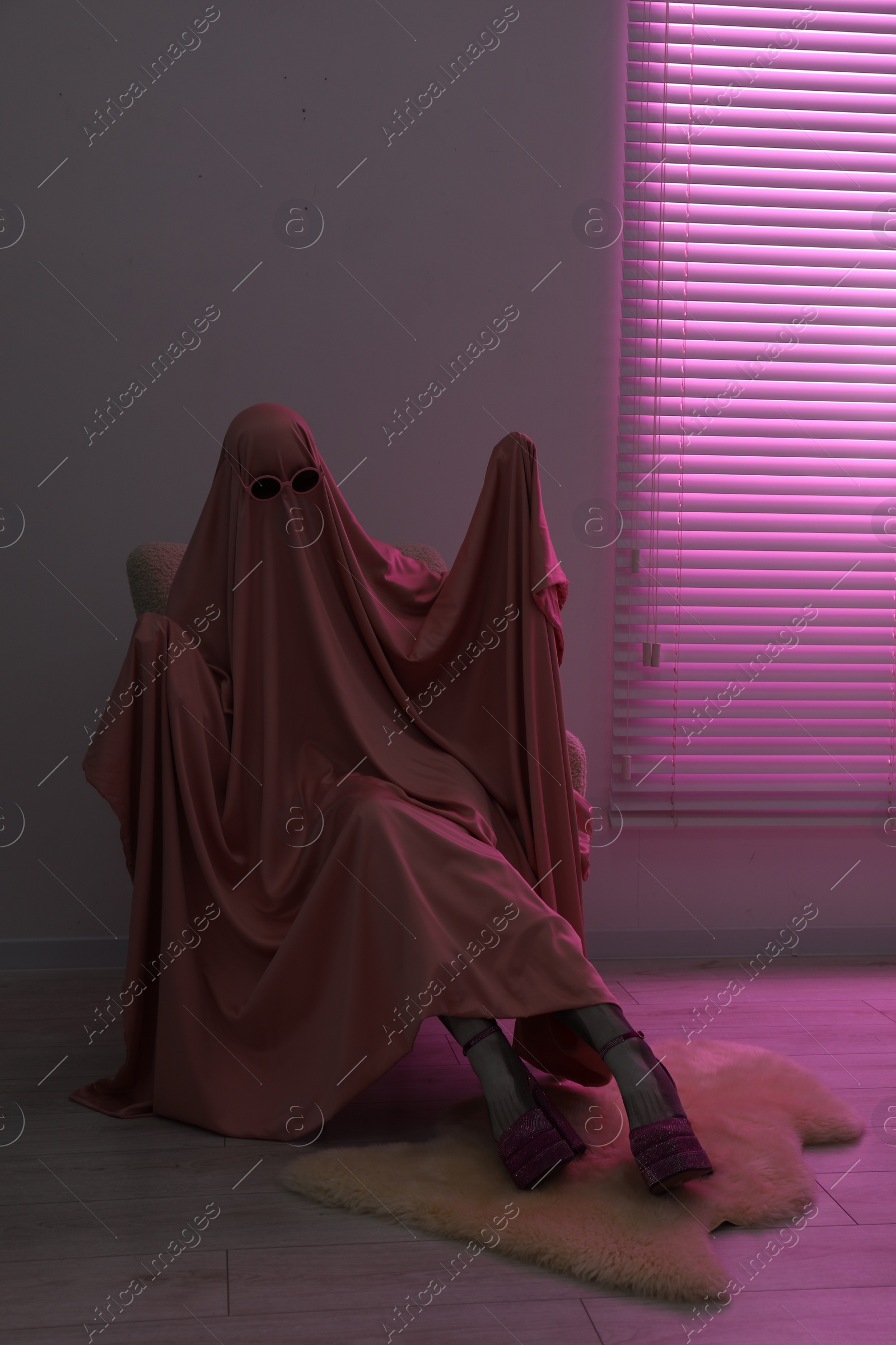 Photo of Glamorous ghost. Woman in sheet and high heel shoes on armchair in pink light