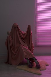 Glamorous ghost. Woman in sheet and high heel shoes on armchair in pink light