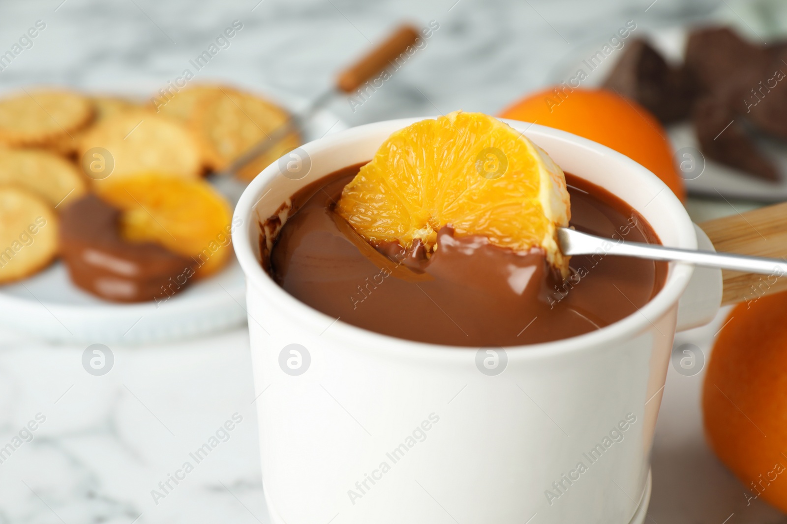 Photo of Dipping orange into fondue pot with chocolate on white marble table, closeup