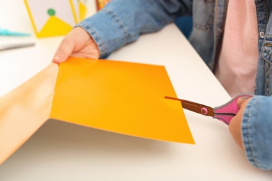 Photo of Little left-handed girl cutting orange construction paper at table, closeup