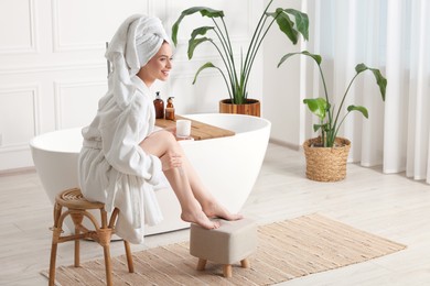 Beautiful young woman applying body cream onto legs in bathroom, space for text