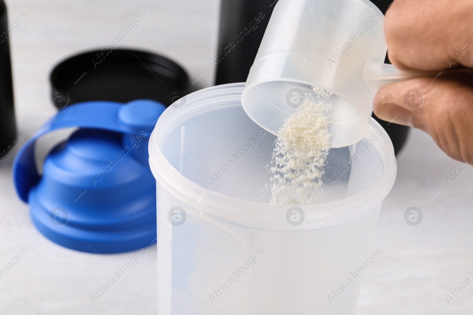 Photo of Man pouring protein powder from measuring scoop into shaker on white table, closeup