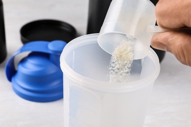Photo of Man pouring protein powder from measuring scoop into shaker on white table, closeup