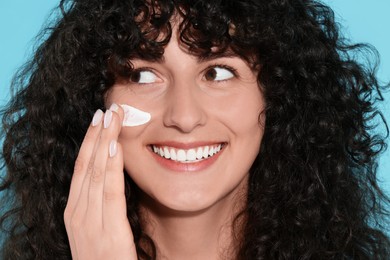 Beautiful young woman applying sun protection cream onto face on light blue background, closeup