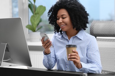 Young woman with cup of drink using smartphone at table in office