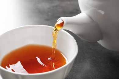 Photo of Pouring black tea into white porcelain cup on gray table, closeup