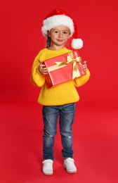 Photo of Cute little girl in Santa hat with Christmas gift on red background