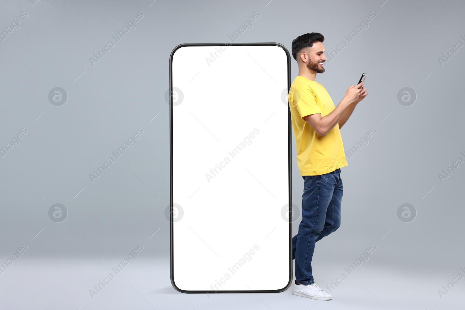 Image of Man with mobile phone standing near huge device with empty screen on grey background. Mockup for design