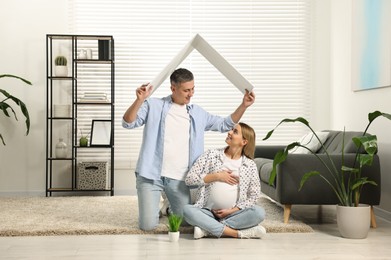 Photo of Young family housing concept. Man holding cardboard roof over his pregnant wife head at home