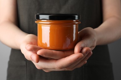 Woman holding glass jar of delicious persimmon jam on gray background, closeup