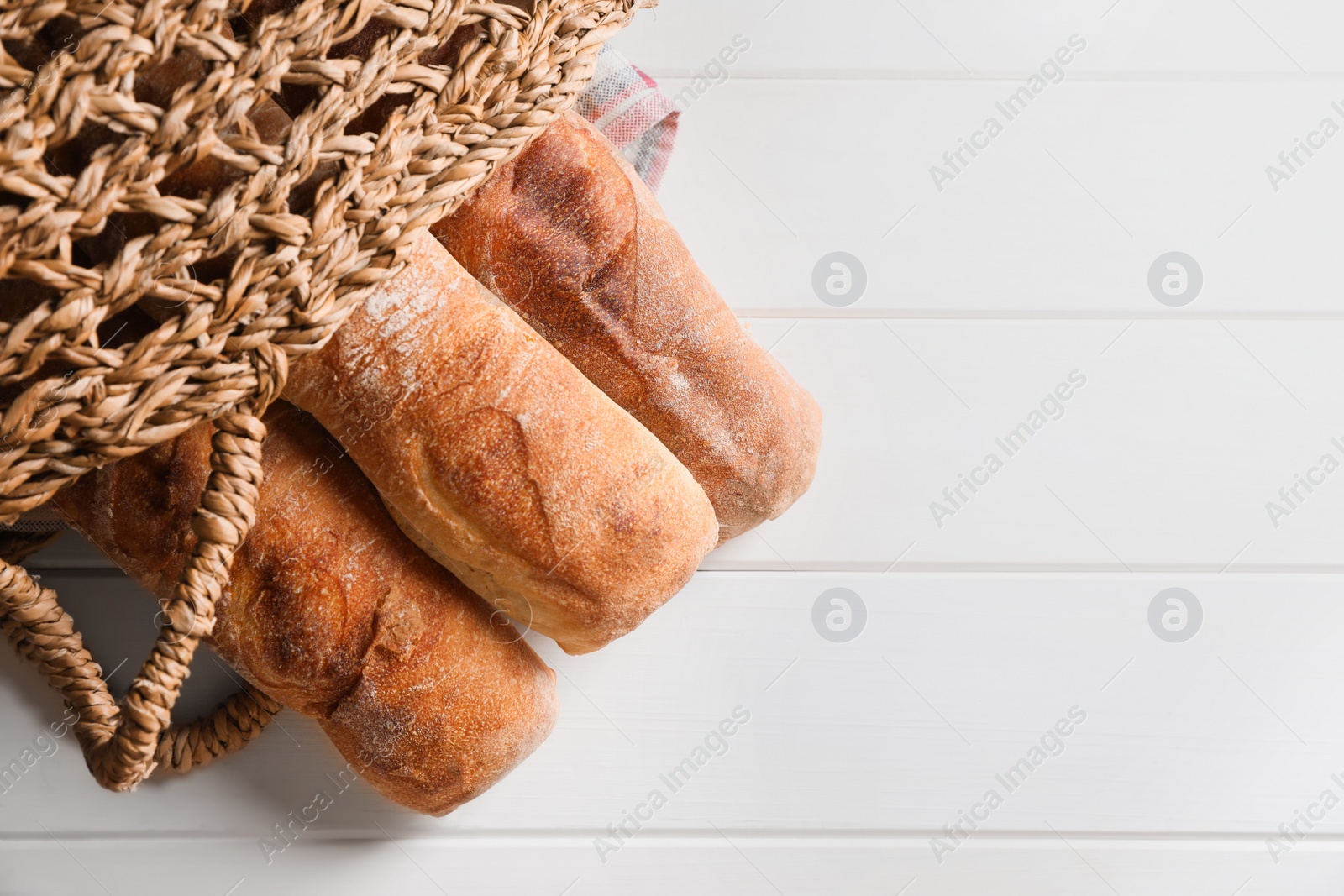 Photo of Wicker bag with fresh crispy ciabattas on white wooden table, top view. Space for text