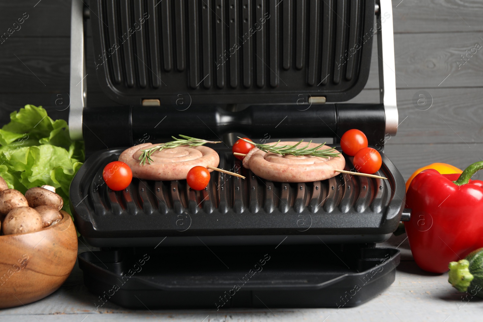 Photo of Electric grill with homemade sausages, rosemary and vegetables on rustic wooden table