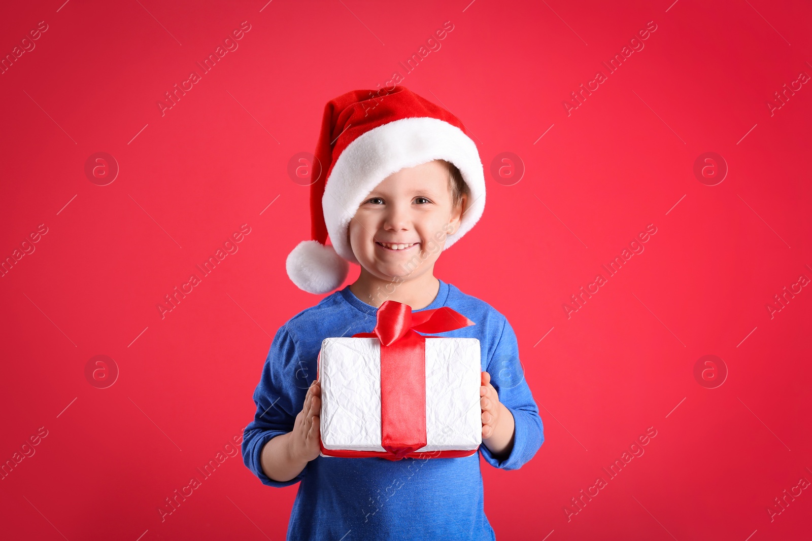 Photo of Cute little boy in Santa Claus hat holding gift box on red background