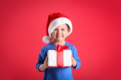 Photo of Cute little boy in Santa Claus hat holding gift box on red background