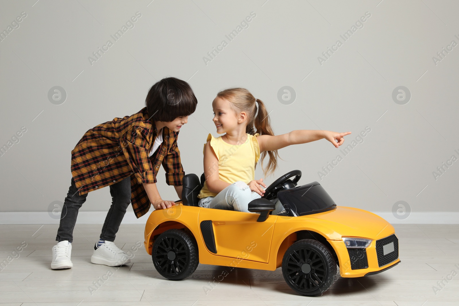 Photo of Cute boy pushing children's electric toy car with little girl near grey wall indoors