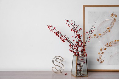 Photo of Hawthorn branches with red berries in vase, picture and decorative letter on wooden table, space for text