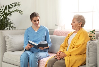 Photo of Nurse reading book to elderly woman indoors. Assisting senior people