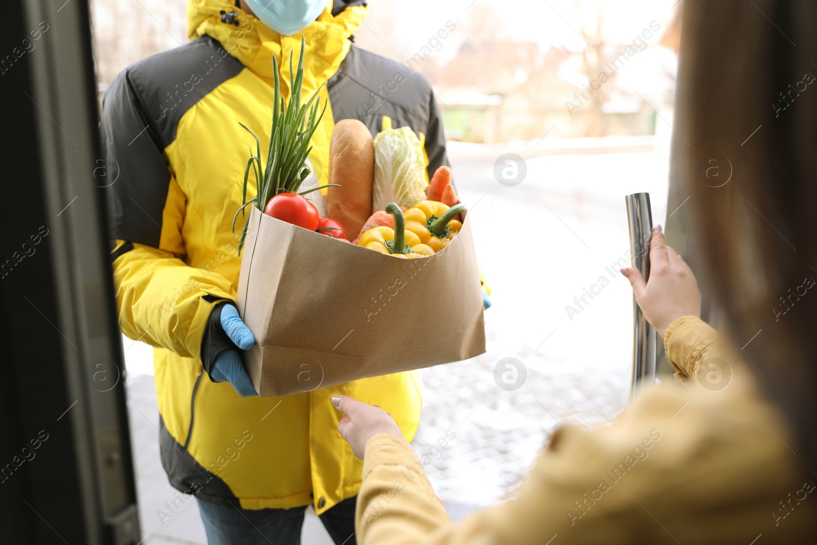 Photo of Courier in medical mask giving paper bag with groceries to woman at doorway, closeup. Delivery service during Covid-19 quarantine