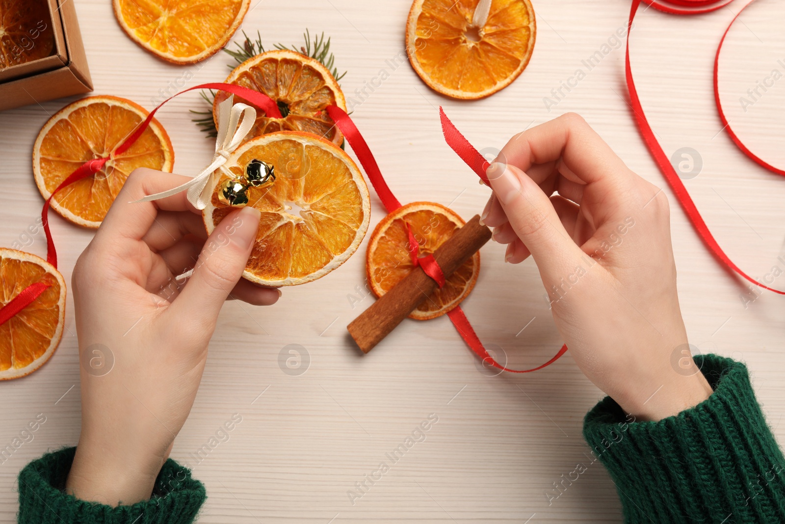 Photo of Woman making garland from dry oranges, cinnamon, ribbon and sleigh bells at white wooden table, top view