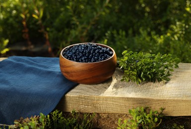 Photo of Delicious bilberries in bowl and branch with fresh berries on wooden bench outdoors