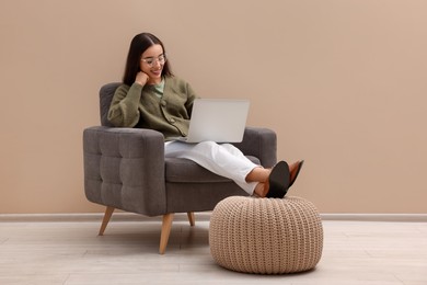 Beautiful woman with laptop sitting in armchair near beige wall indoors