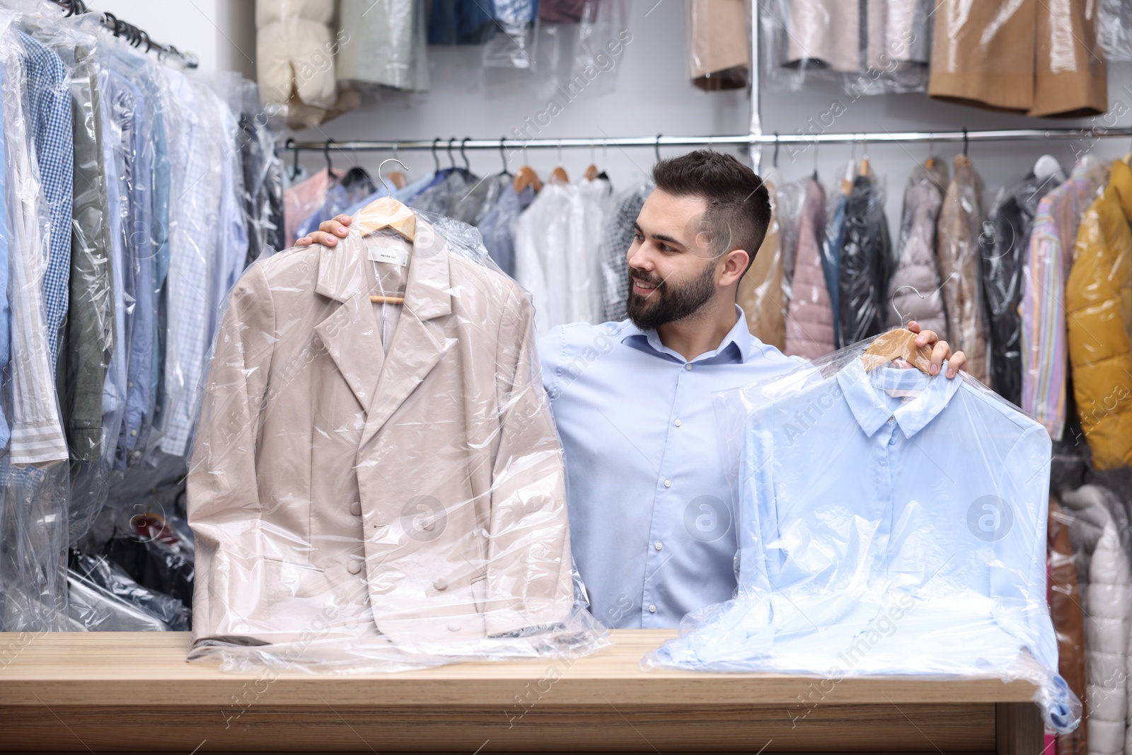 Photo of Dry-cleaning service. Happy worker holding hangers with clothes in plastic bags at counter indoors