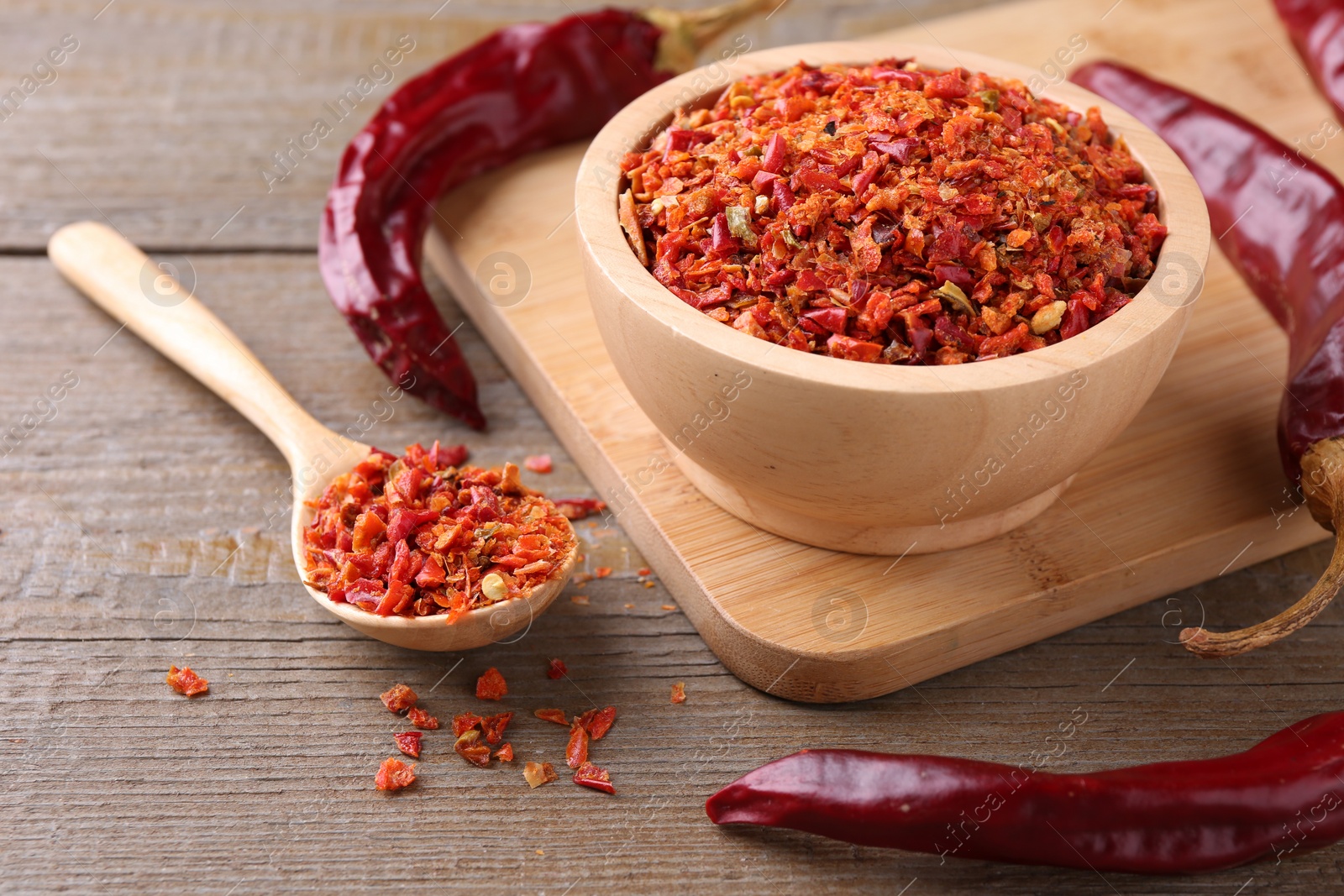 Photo of Aromatic spice. Red chili pepper flakes in bowl, spoon and pods on wooden table, closeup