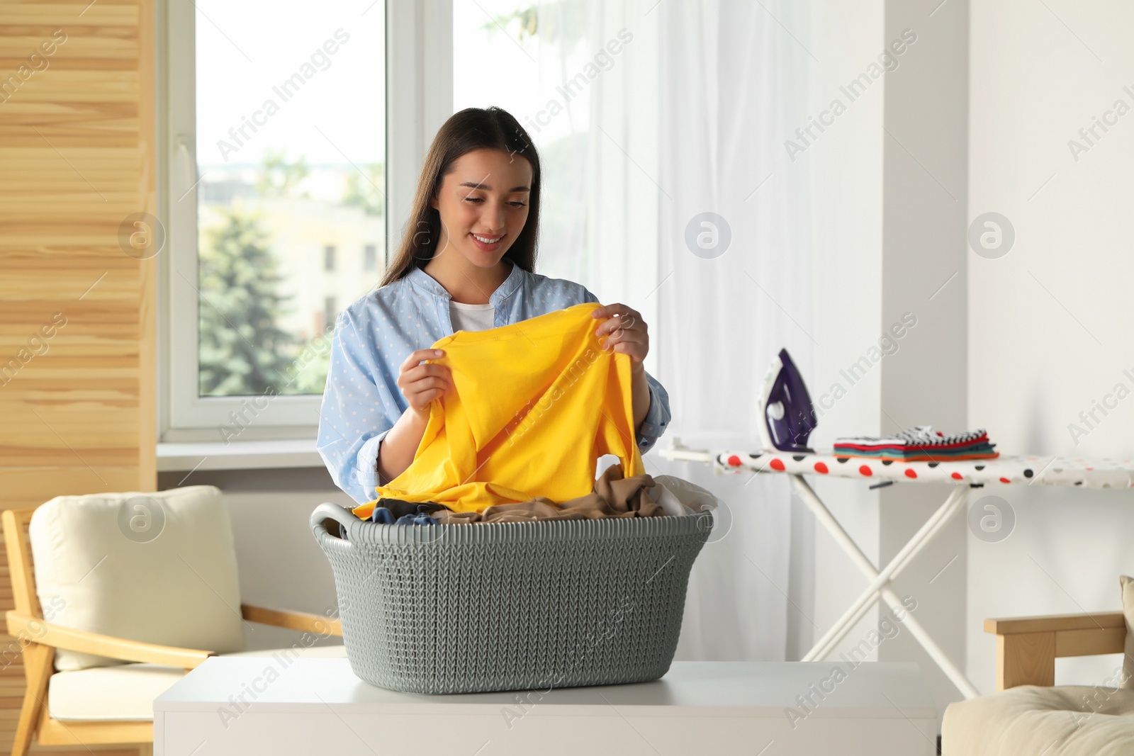 Photo of Young woman with basket full of clean laundry at table indoors