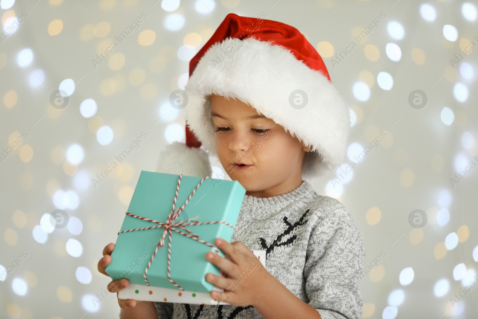 Photo of Surprised little child in Santa hat with gift box against blurred festive lights. Christmas celebration