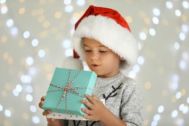 Surprised little child in Santa hat with gift box against blurred festive lights. Christmas celebration