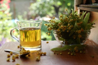 Photo of Glass cup of aromatic tea, spoon and linden blossoms on wooden table against blurred background