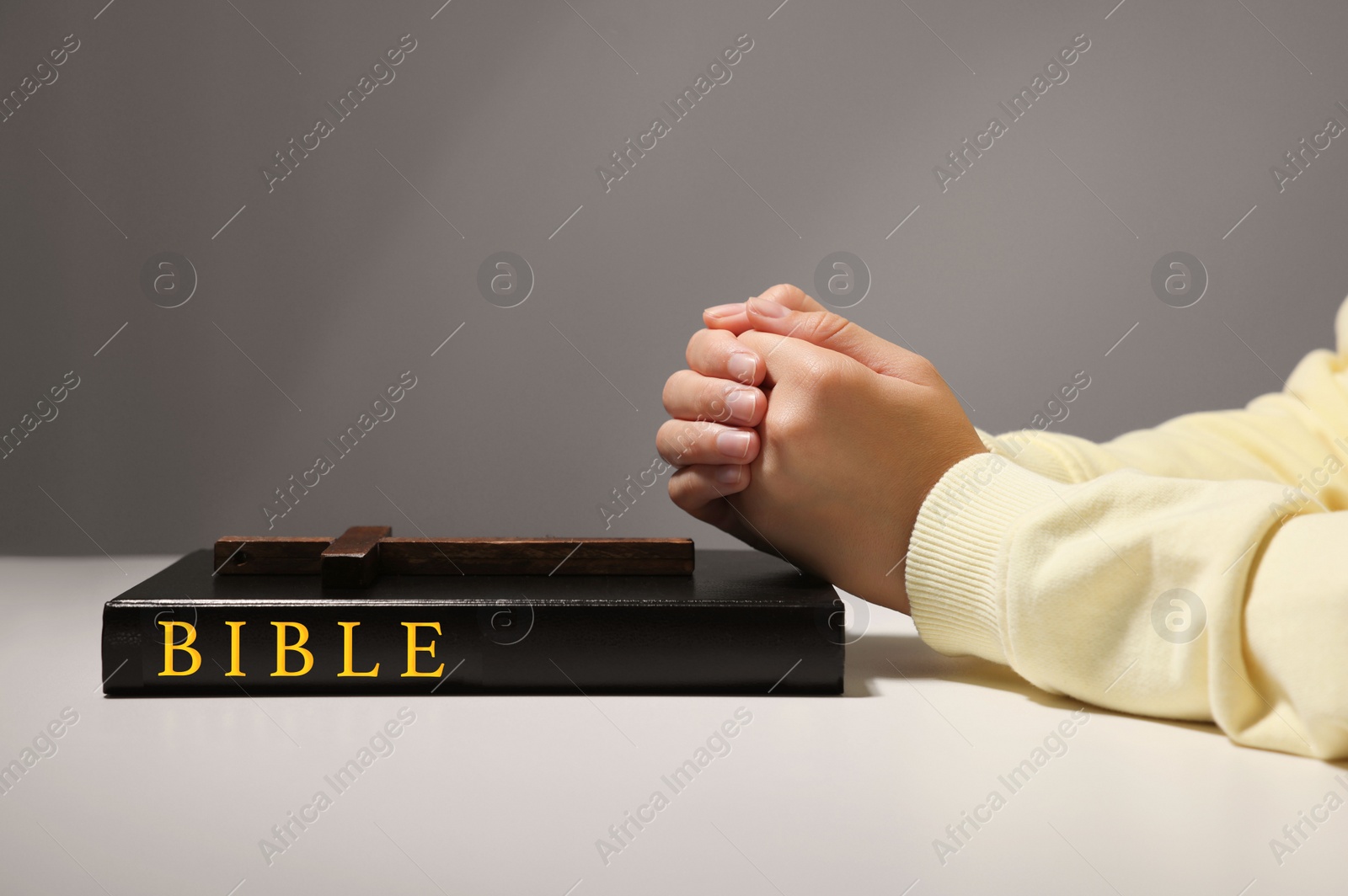 Photo of Woman praying over Bible with wooden cross at white table against grey background, closeup