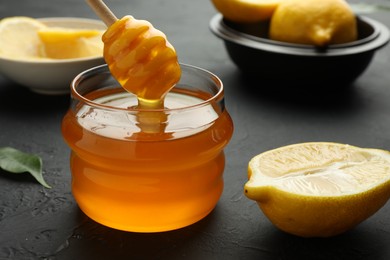 Dripping sweet honey from dipper into jar and fresh lemons on black table, closeup