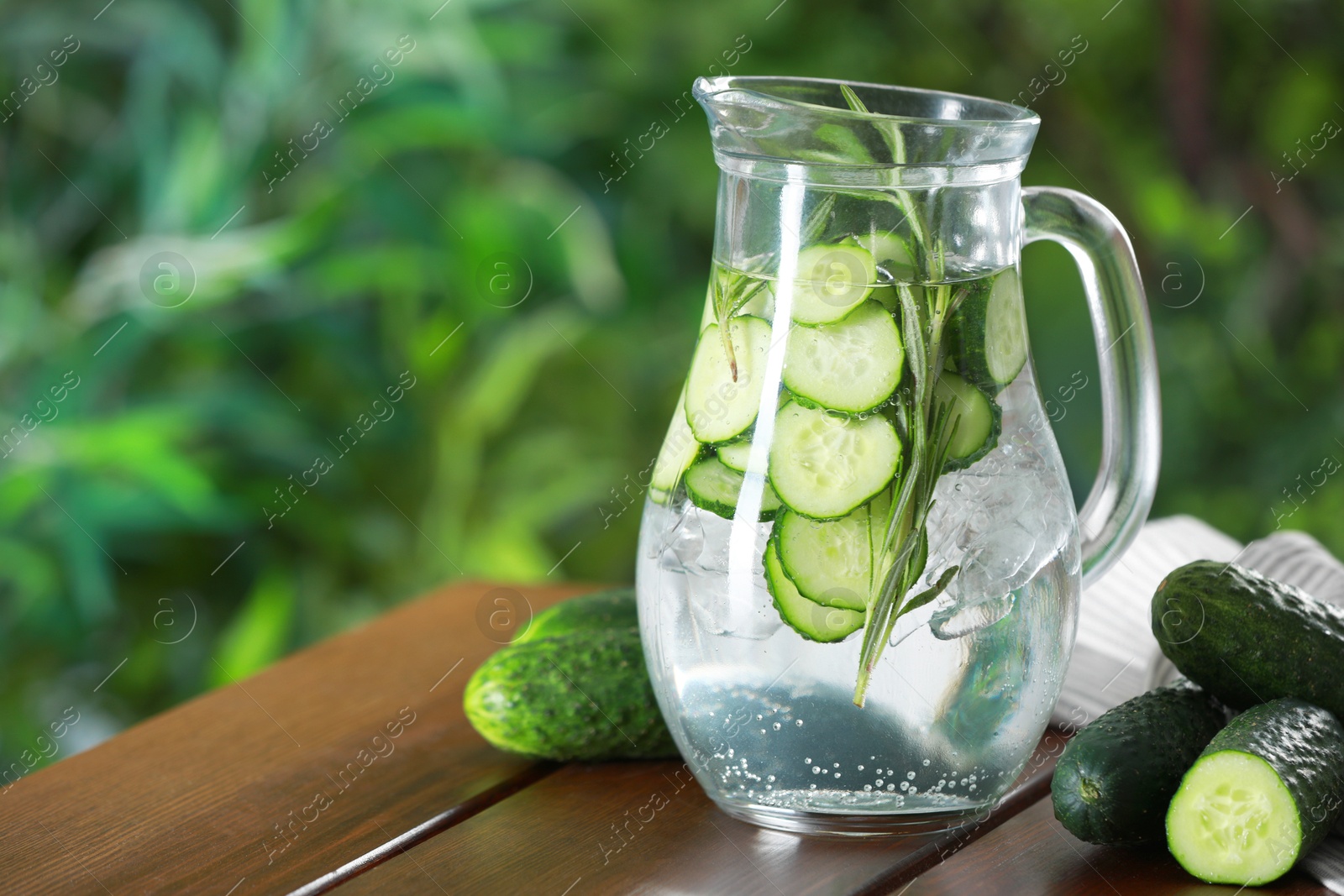 Photo of Refreshing cucumber water with rosemary in jug and vegetables on wooden table against blurred green background, closeup. Space for text