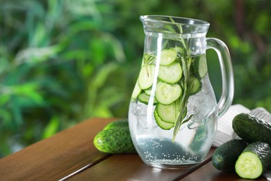 Photo of Refreshing cucumber water with rosemary in jug and vegetables on wooden table against blurred green background, closeup. Space for text