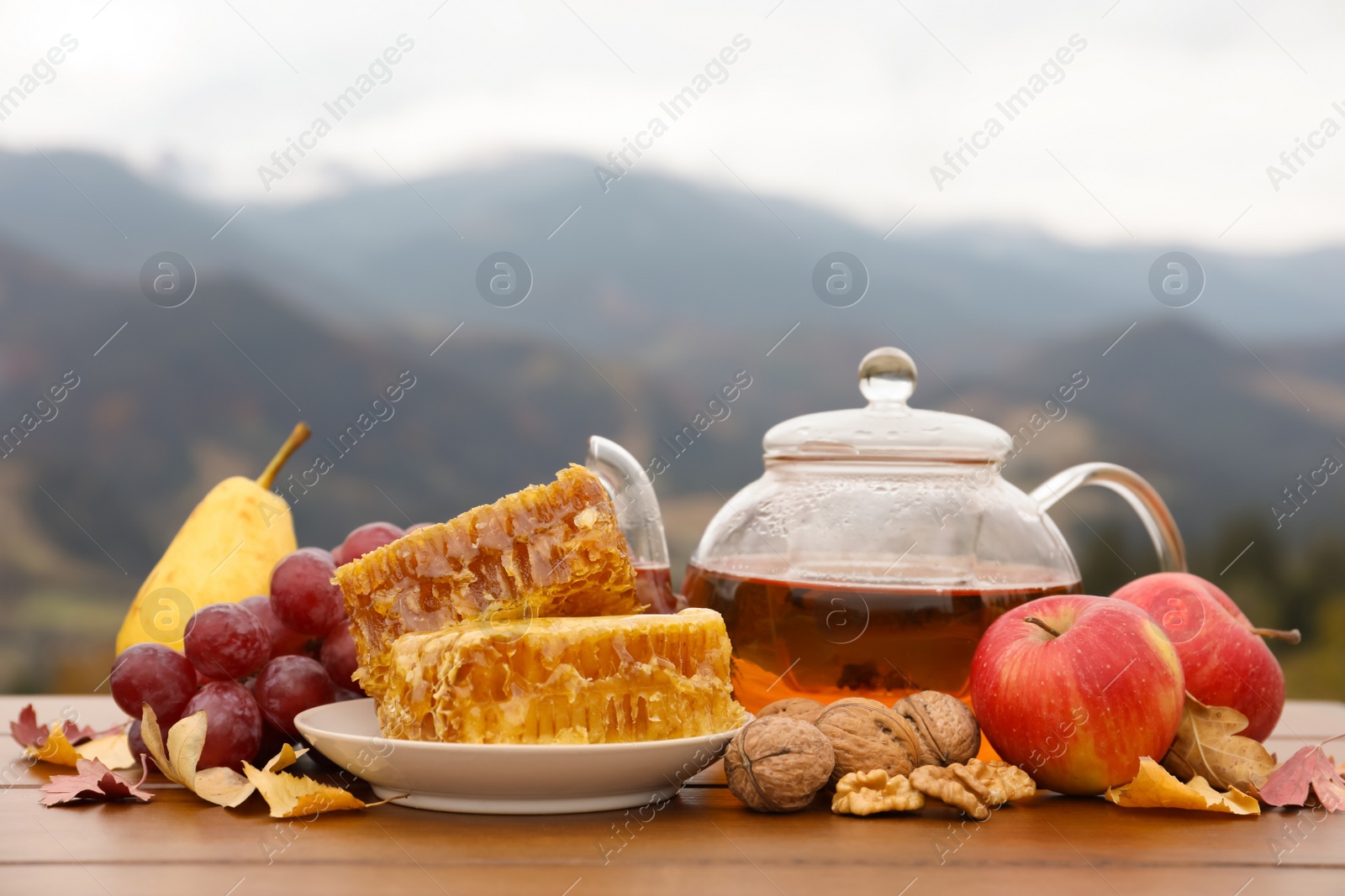 Photo of Tasty fresh honeycombs, fruits, walnuts and aromatic tea on wooden table against mountain landscape