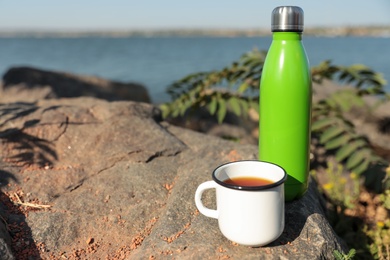 Photo of Modern green thermos bottle and cup on stone near river. Space for text