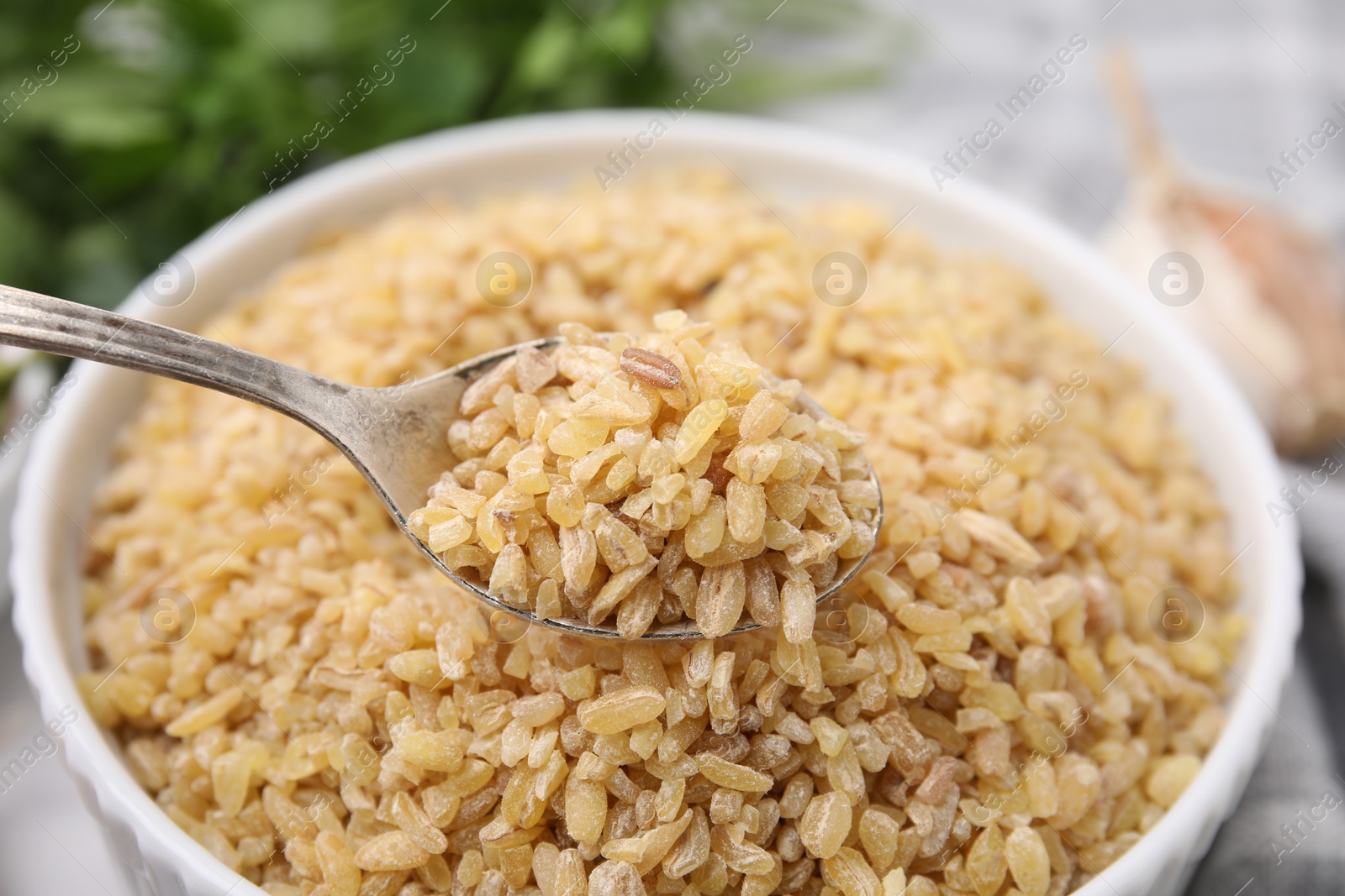 Photo of Taking raw bulgur with spoon from bowl on table, closeup