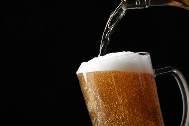 Photo of Pouring cold tasty beer from bottle into glass mug on black background, closeup. Space for text