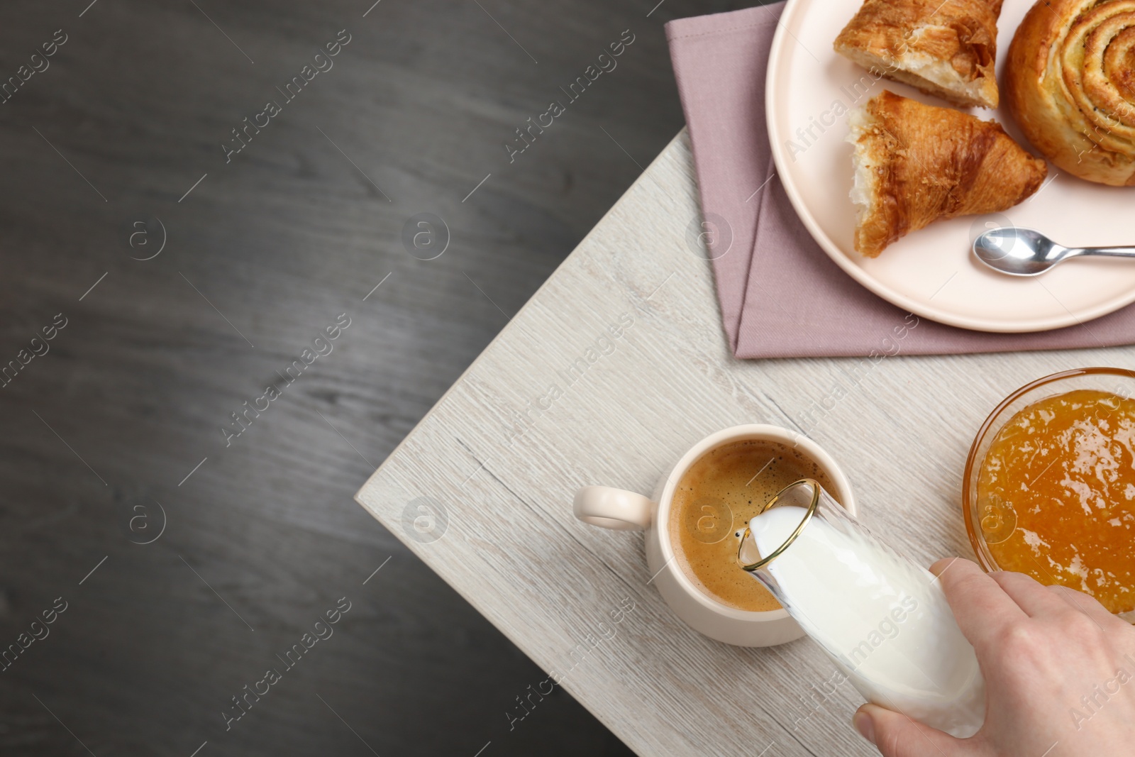 Photo of Woman pouring milk into cup with coffee and pastry at wooden table, top view. space for text