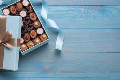 Open box of delicious chocolate candies and color ribbon on light blue wooden table, flat lay. Space for text