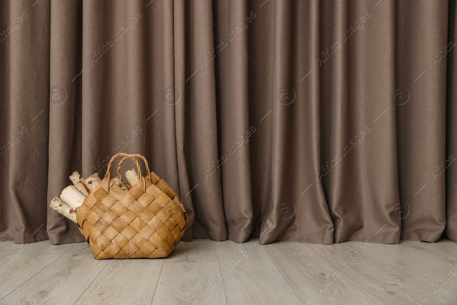 Photo of Wicker bag with firewood on floor near elegant curtains indoors, space for text