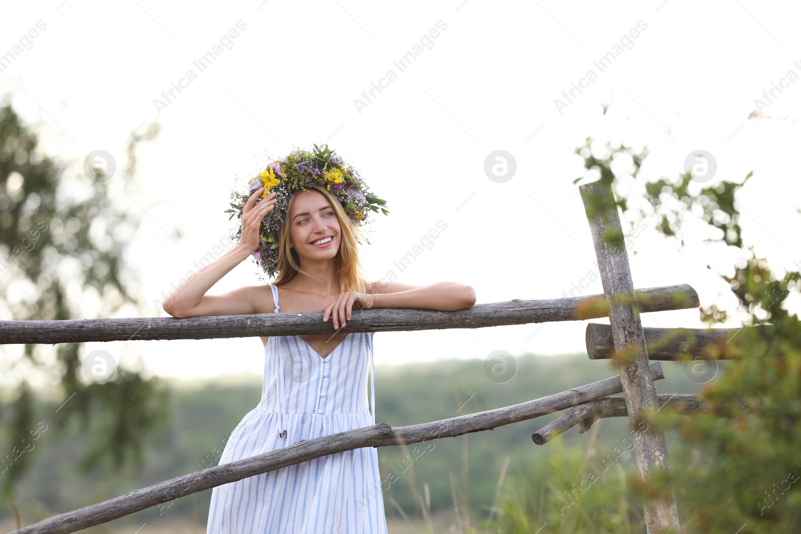 Photo of Young woman wearing wreath made of beautiful flowers near wooden fence